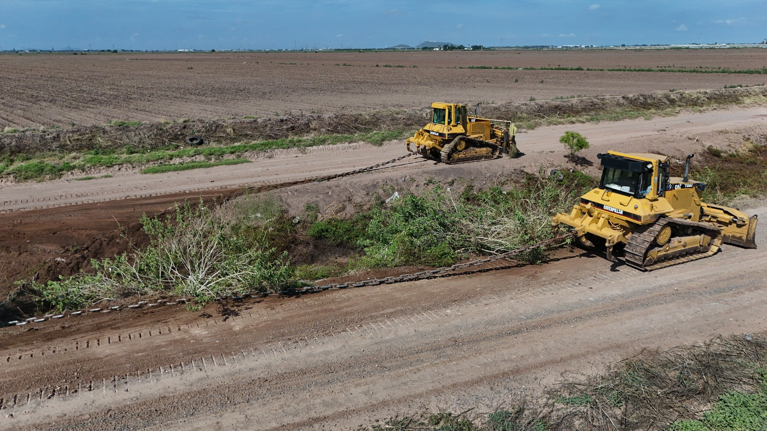 Desazolve en cadena · dos bulldozers. Dos Caterpillar D6 arrastran cadena sobre el bordo del canal para retirar vegetación. Toma aérea de 36 MP.